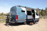 An RV rental camper van parked in nature. A person relaxes under the awning, another handles the bike rack at the back.