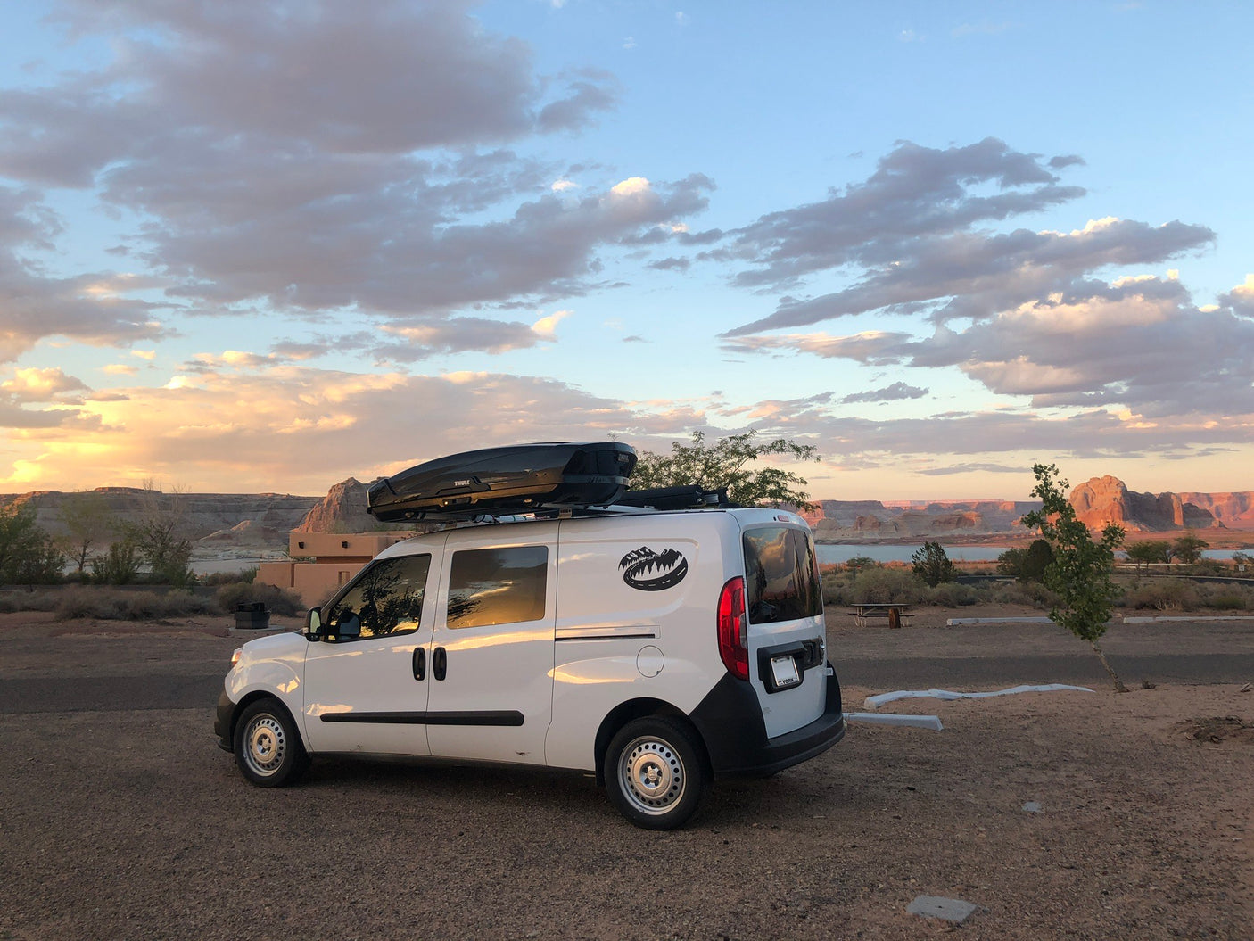 Compact white RV rental camper van parked at sunset beside a desert lake, framed by colorful clouds. Ready for adventure.