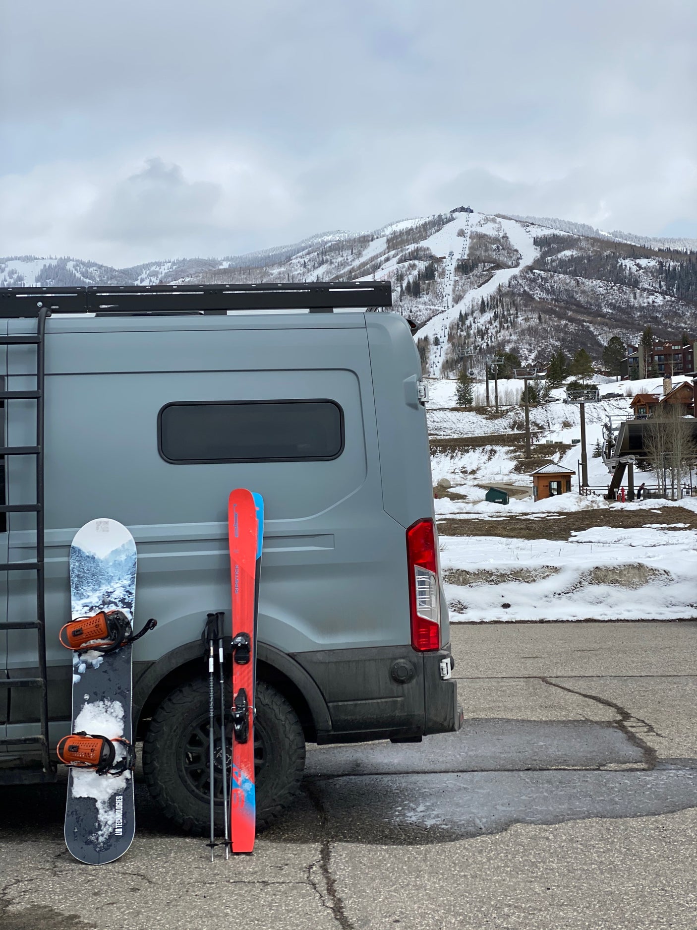 RV rental ready for ski day. Grey van with snowboard & skis, snow-covered mountains, & ski lifts in background.