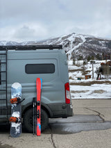 RV rental ready for ski day. Grey van with snowboard & skis, snow-covered mountains, & ski lifts in background.
