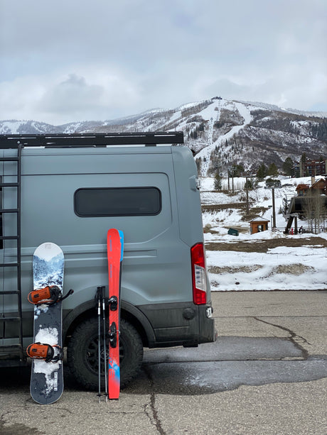 RV rental ready for ski day. Grey van with snowboard & skis, snow-covered mountains, & ski lifts in background.