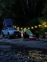 Nighttime riverside camping: truck with rooftop tent, ground tent, string lights. A comfy setup for your next rv rental adventure.