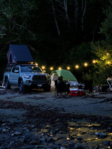 Nighttime riverside camping: truck with rooftop tent, ground tent, string lights. A comfy setup for your next rv rental adventure.
