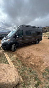 Dark gray rv rental camper van parked on a dirt lot with mountains in distance under cloudy skies.