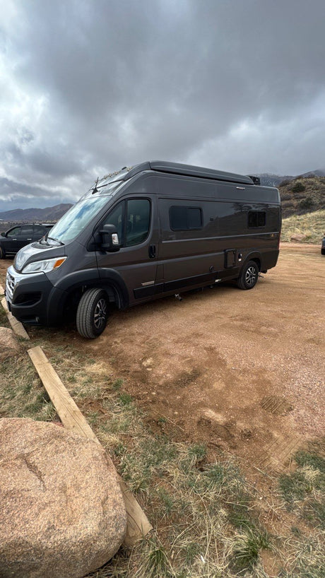Dark gray rv rental camper van parked on a dirt lot with mountains in distance under cloudy skies.