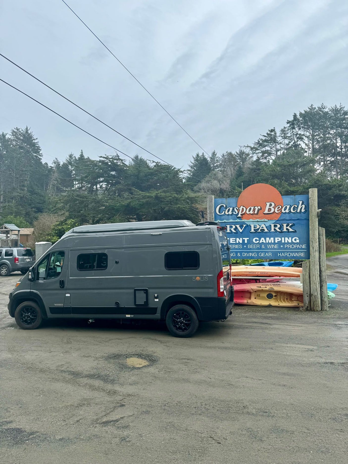 A gray rv rental van parked at the Caspar Beach RV Park sign, listing camping, groceries, propane, and kayaks.