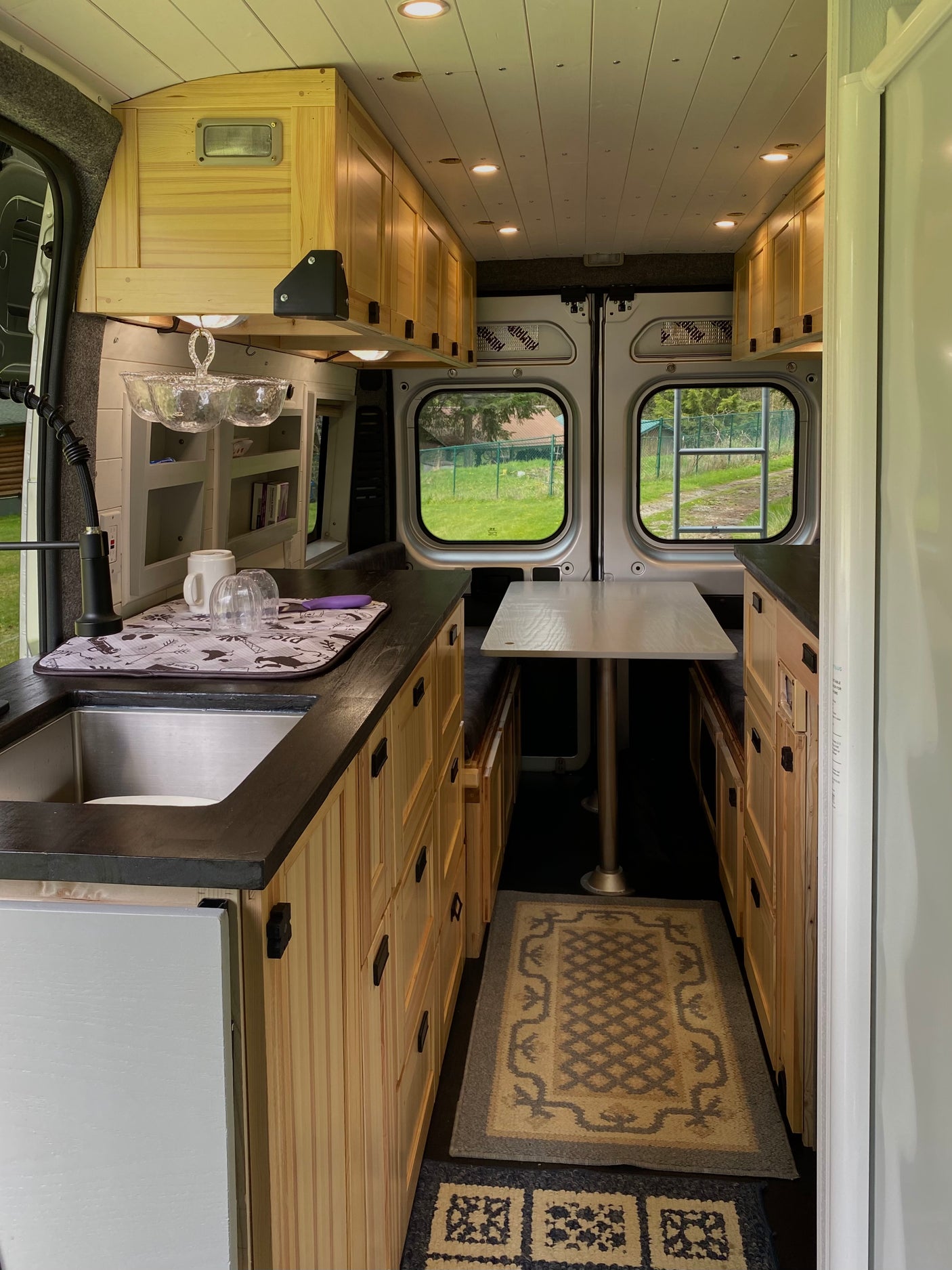 Spacious RV rental interior: kitchen with sink, natural wood cabinets, dining table, and large windows revealing green outdoor scenery.