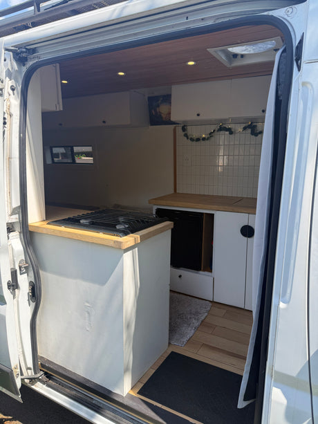 View into an rv rental camper van's modern kitchen with stove & cabinets, plus a bed platform, from the open side door.