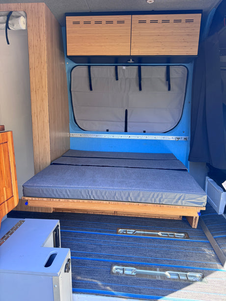 Ready for adventure? This RV rental offers a compact camper van interior with a bed, wood cabinets, and blue accents.
