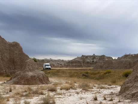 An RV rental van navigates a rugged Badlands landscape of eroded hills and dry grass under a cloudy sky.