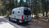 RV rental: A light gray Winnebago Revel camper van, with roof rack and rear ladder, parked by a pine forest.