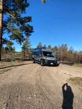 A grey rv rental campervan on a gravel road, surrounded by pine trees, some burnt, under a clear blue sky.