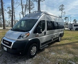 Silver Roadtrek camper van RV rental parked on grass in a sunny, tree-filled campground.