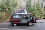 Red pop-top camper van RV rental parked in forest campsite. Side door open, interior visible.