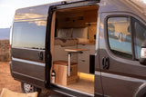 Explore this cozy gray camper van interior, available for rv rental. Bed, table, & wood paneling in a desert setting.