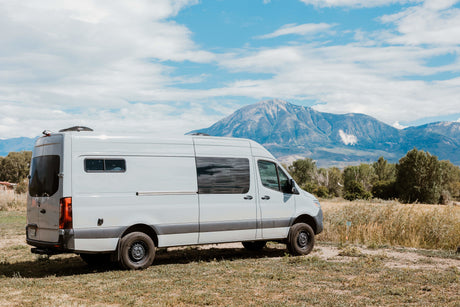 White RV rental van parked in a field with a majestic mountain and cloudy blue sky backdrop. Ideal for adventure travel.