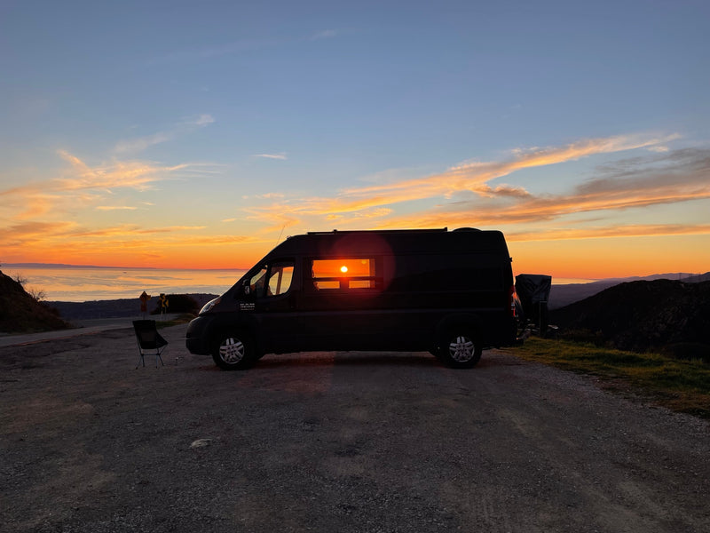 A black RV rental faces a breathtaking sunset over the ocean from a scenic overlook, with the sun radiating through its side window. — photo 3