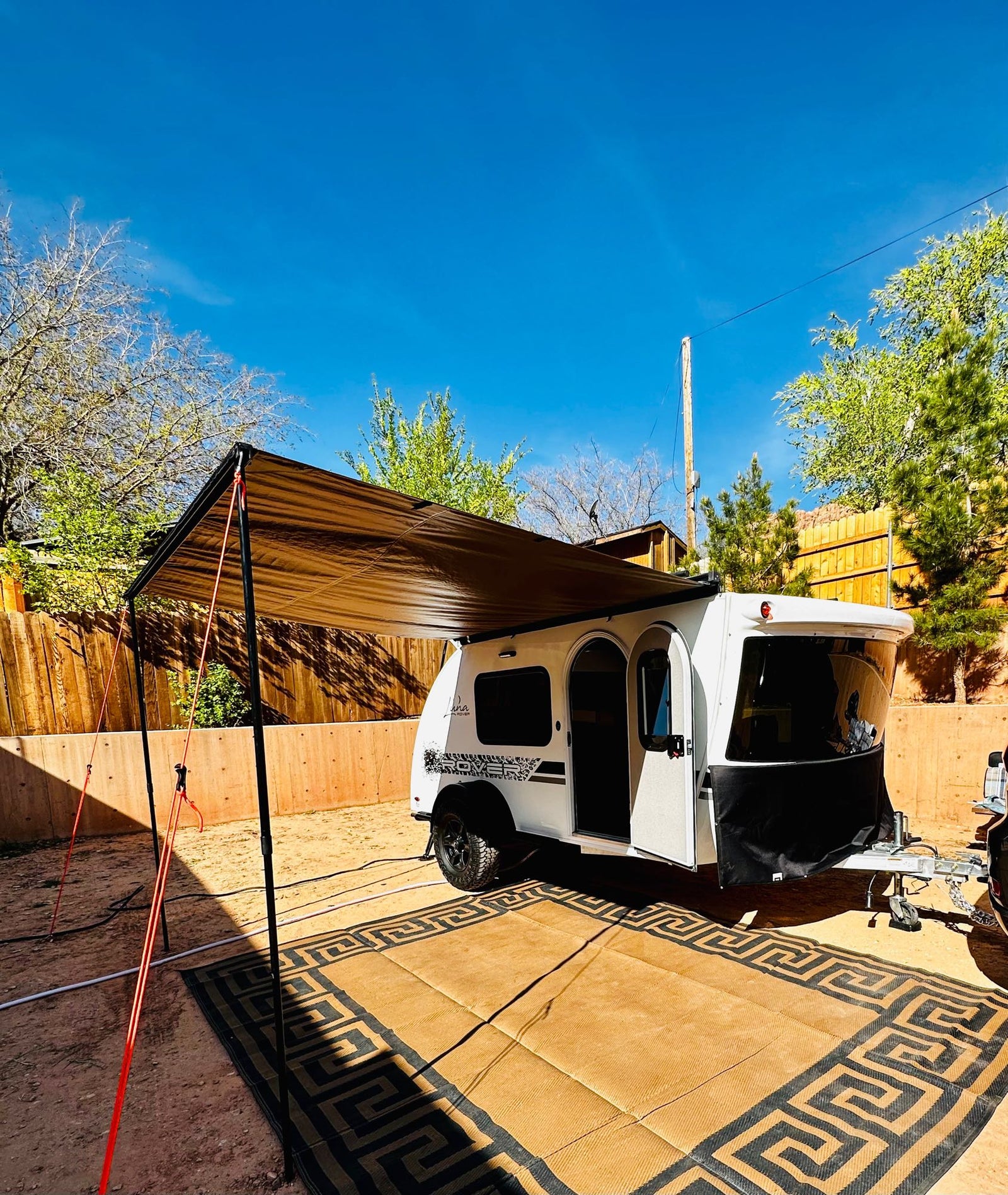 White teardrop RV rental showcasing its setup with a brown awning and patterned outdoor mat at a campsite.