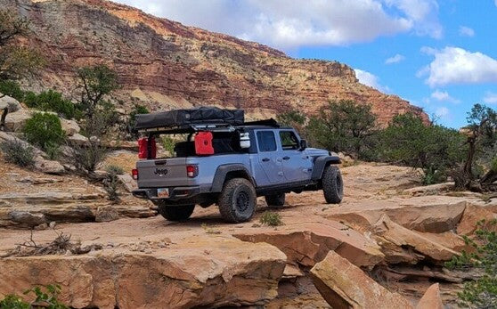 A modified Jeep Gladiator pickup with a rooftop tent on a rocky trail, red desert mountains behind. Ideal off-road RV rental adventure.