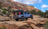 A modified Jeep Gladiator pickup with a rooftop tent on a rocky trail, red desert mountains behind. Ideal off-road RV rental adventure.