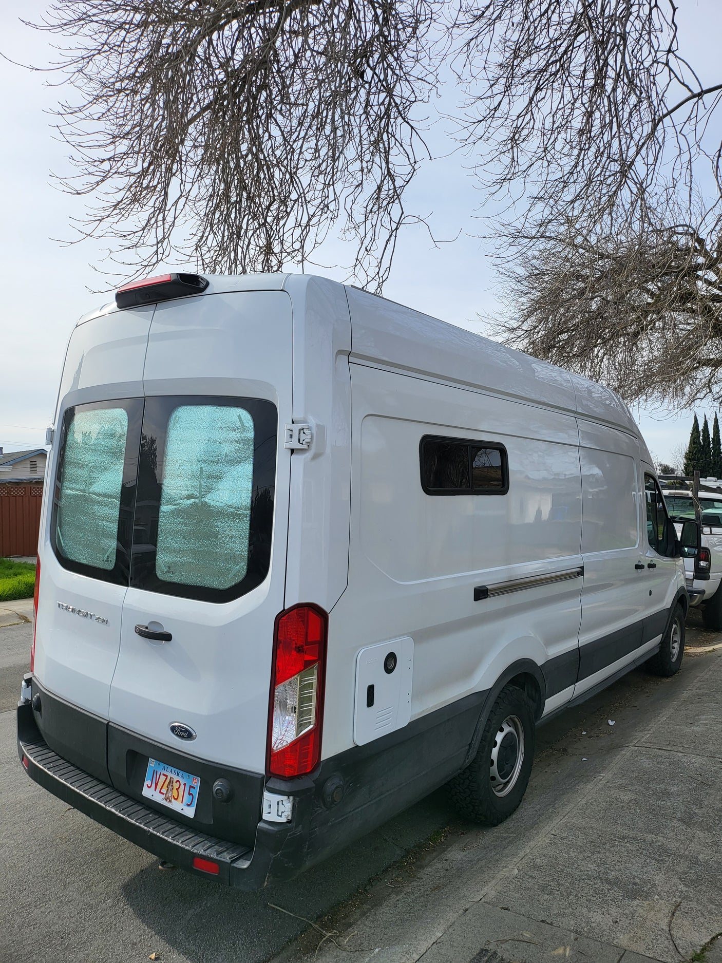 White Ford Transit camper van, an RV rental, parked with Alaska plates. Bare trees above.