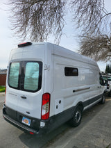 White Ford Transit camper van, an RV rental, parked with Alaska plates. Bare trees above.