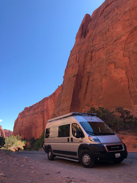 A silver RV rental is parked on a rugged dirt road alongside immense red rock canyon cliffs, under a bright blue sky.