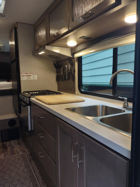 View of a compact RV rental kitchen with double sink, stove, cabinets, and hanging utensils. Functional design for travel.