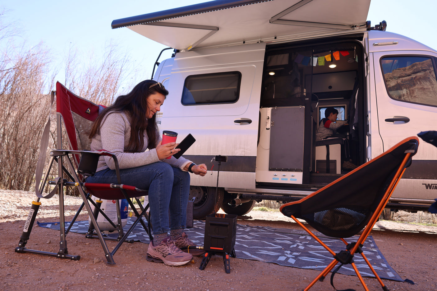 Enjoying an RV rental: a woman uses her tablet outside while a child relaxes inside the camper van, perfect for outdoor living.