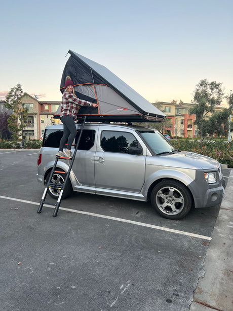 A woman sets up a Roofnest rooftop tent on a silver Honda Element, perfect for unique RV rental camping adventures.