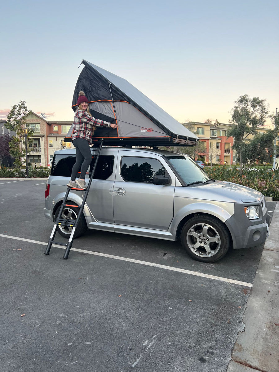A woman sets up a Roofnest rooftop tent on a silver Honda Element, perfect for unique RV rental camping adventures.