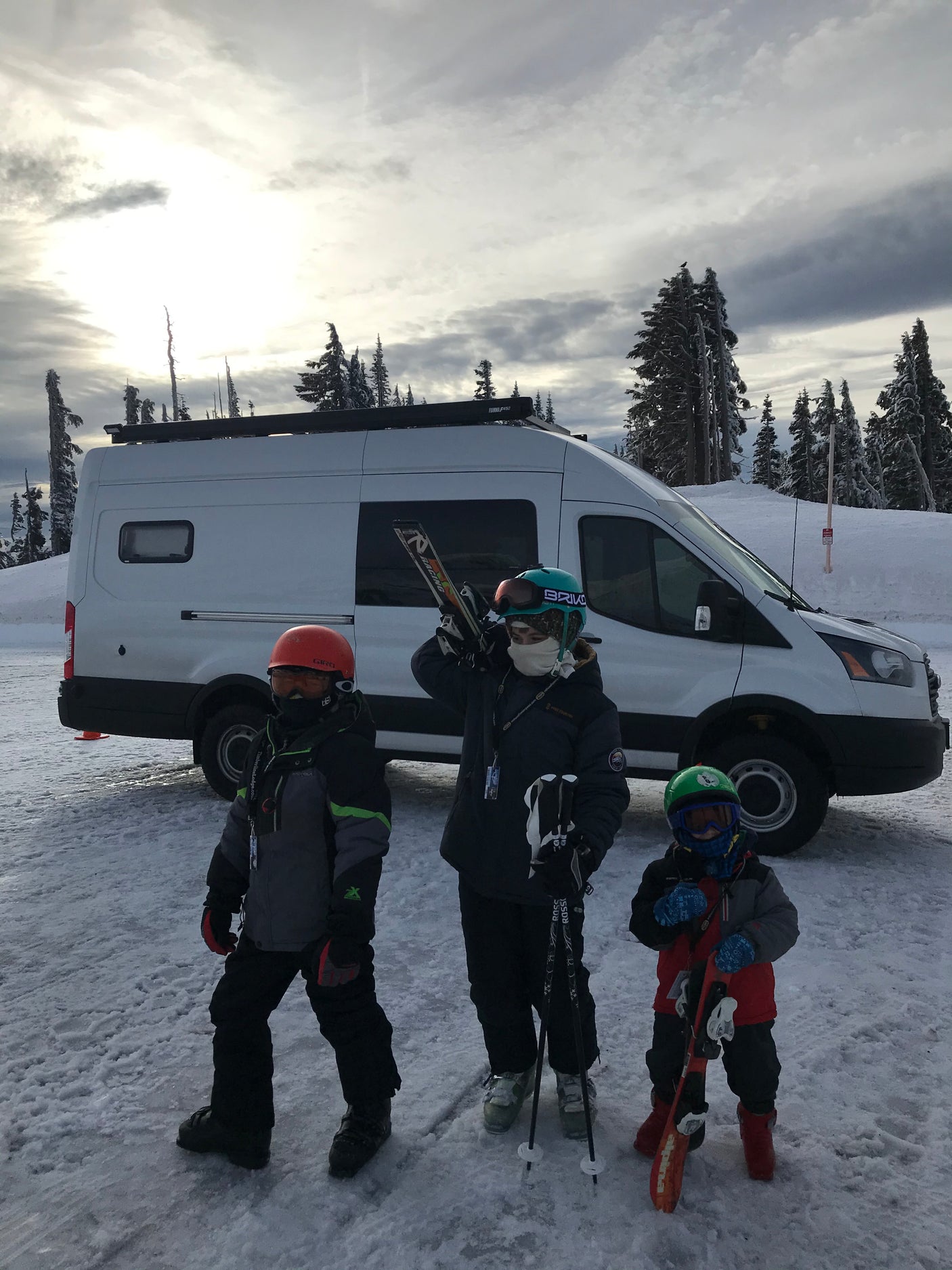 Three young skiers, dressed in colorful gear, stand by their white camper van RV rental in a snowy mountain resort.