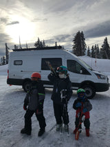 Three young skiers, dressed in colorful gear, stand by their white camper van RV rental in a snowy mountain resort.