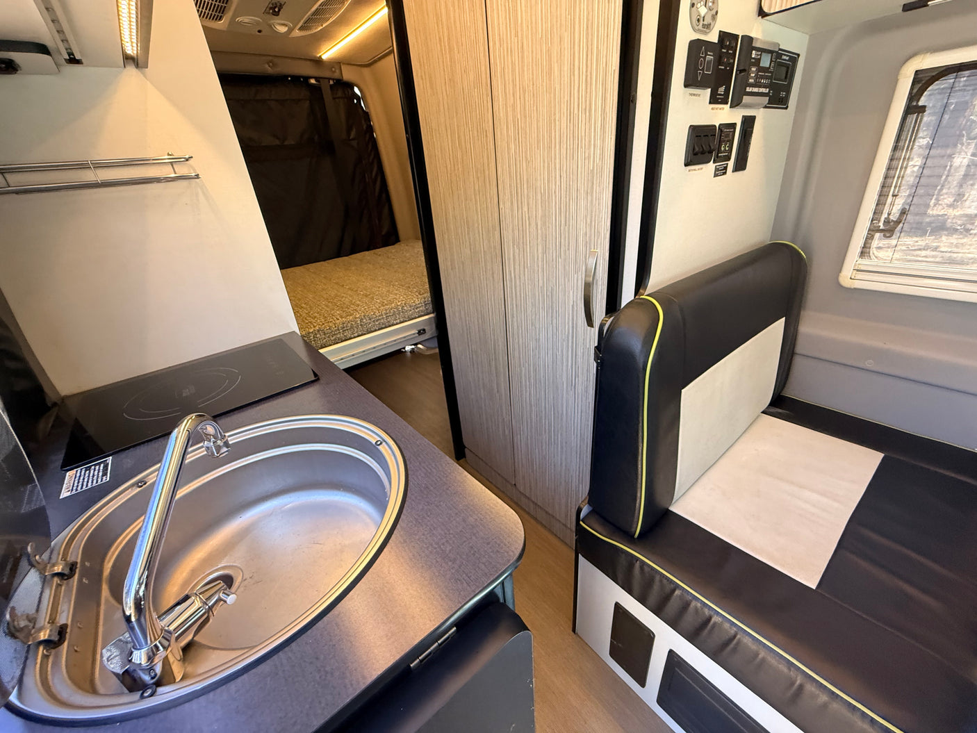 Compact rv rental interior: kitchenette with sink & stovetop, next to a black & white sofa. A bed is visible in the back.