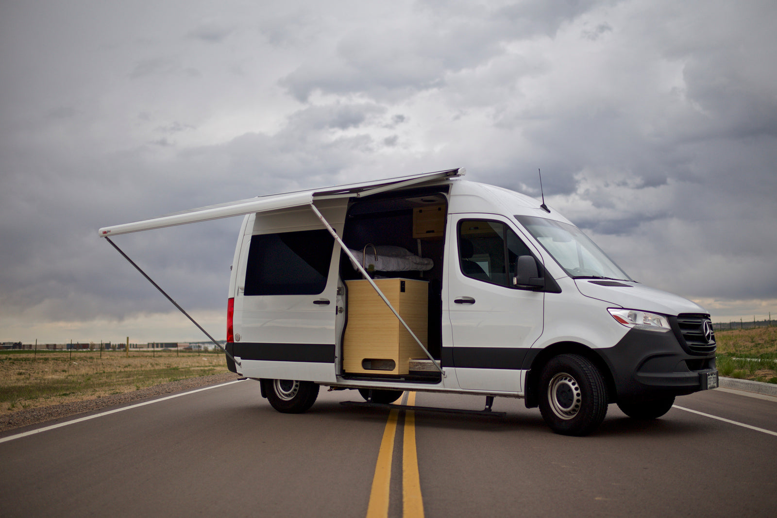 White Mercedes Sprinter RV rental with open side door, custom interior, and extended awning on a road. Ready for adventure!