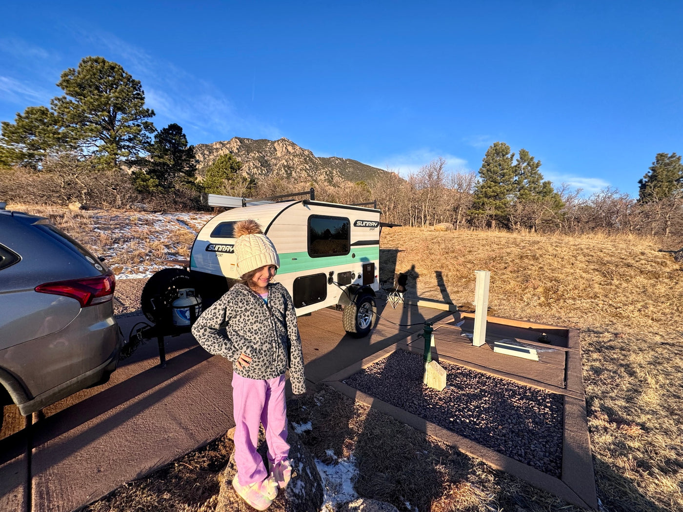 Young girl smiles by a white & teal Sunray Sport RV rental teardrop trailer at a sunny mountain campsite. — photo 9