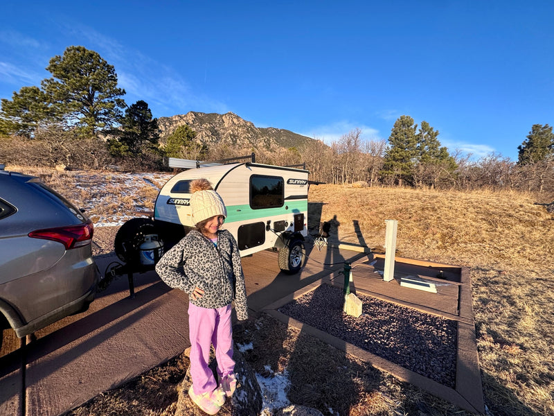Young girl smiles by a white & teal Sunray Sport RV rental teardrop trailer at a sunny mountain campsite. — photo 9