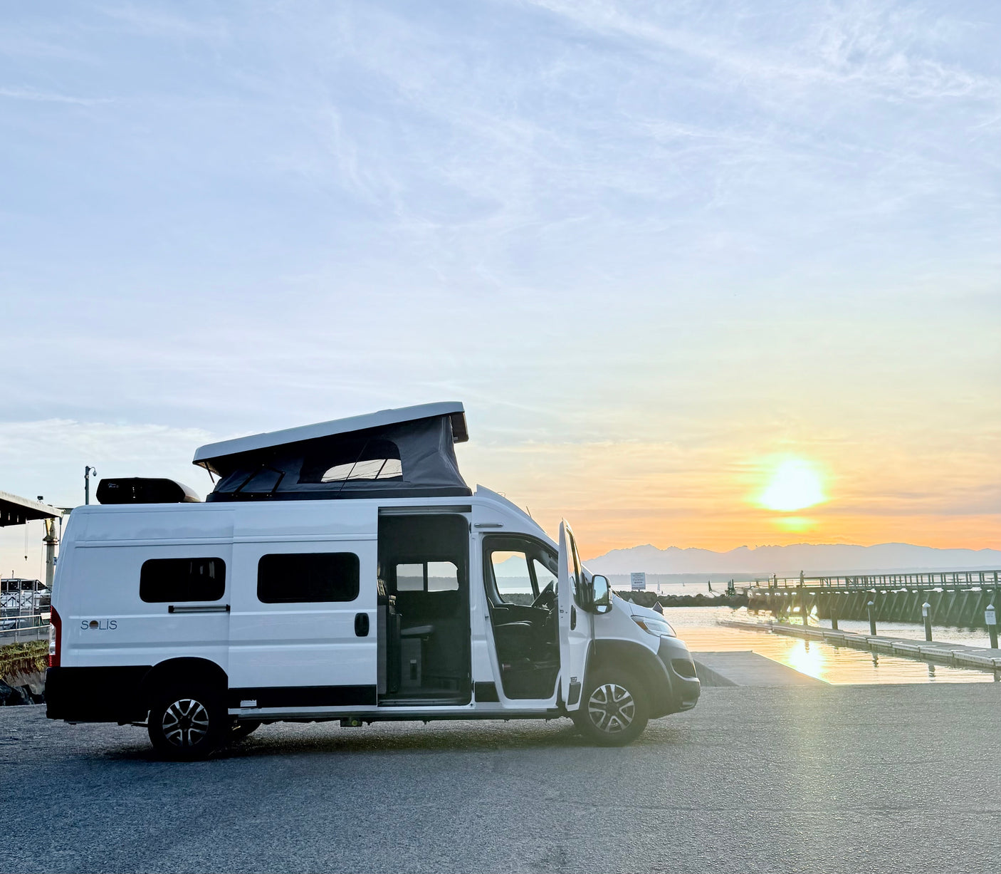 Experience an **rv rental** adventure! A white camper van with its pop-top extended, parked at sunset by a waterfront and distant mountains.