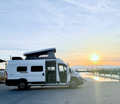 Experience an **rv rental** adventure! A white camper van with its pop-top extended, parked at sunset by a waterfront and distant mountains.