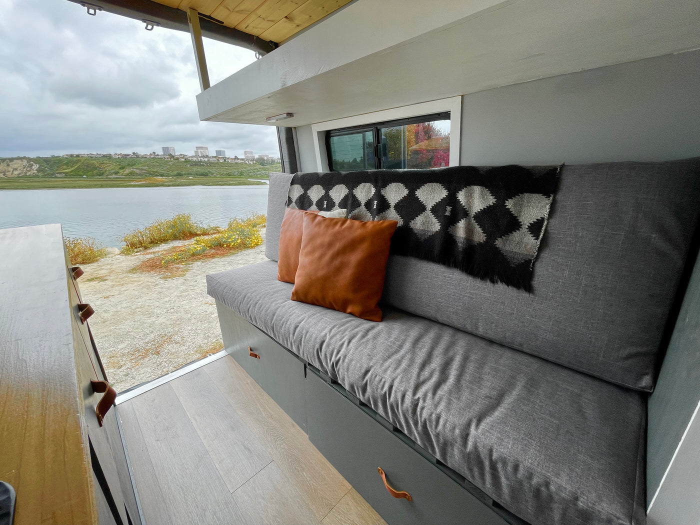 Cozy RV rental interior: gray sofa, brown pillows, patterned blanket, and a large window framing a lake view. — photo 14
