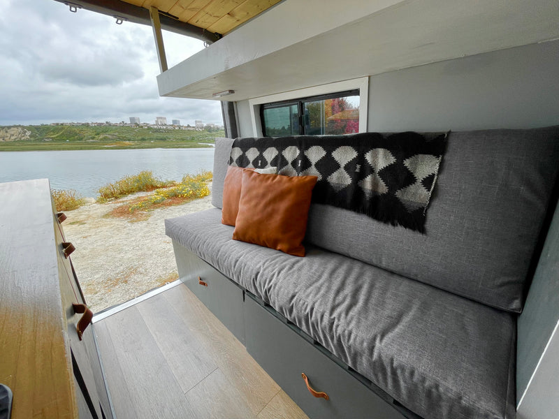 Cozy RV rental interior: gray sofa, brown pillows, patterned blanket, and a large window framing a lake view. — photo 14