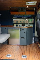 RV rental interior: compact kitchen with sink, stainless fridge, wood counters, and a light grey bench seat.