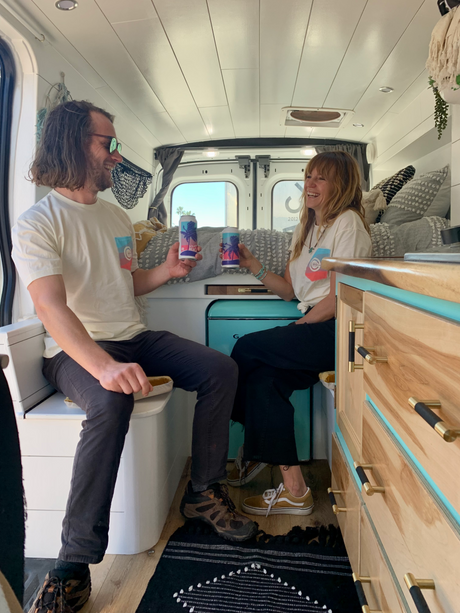 Two people share a laugh inside a cozy rv rental, holding cans with palm trees. The van has a modern wood interior.