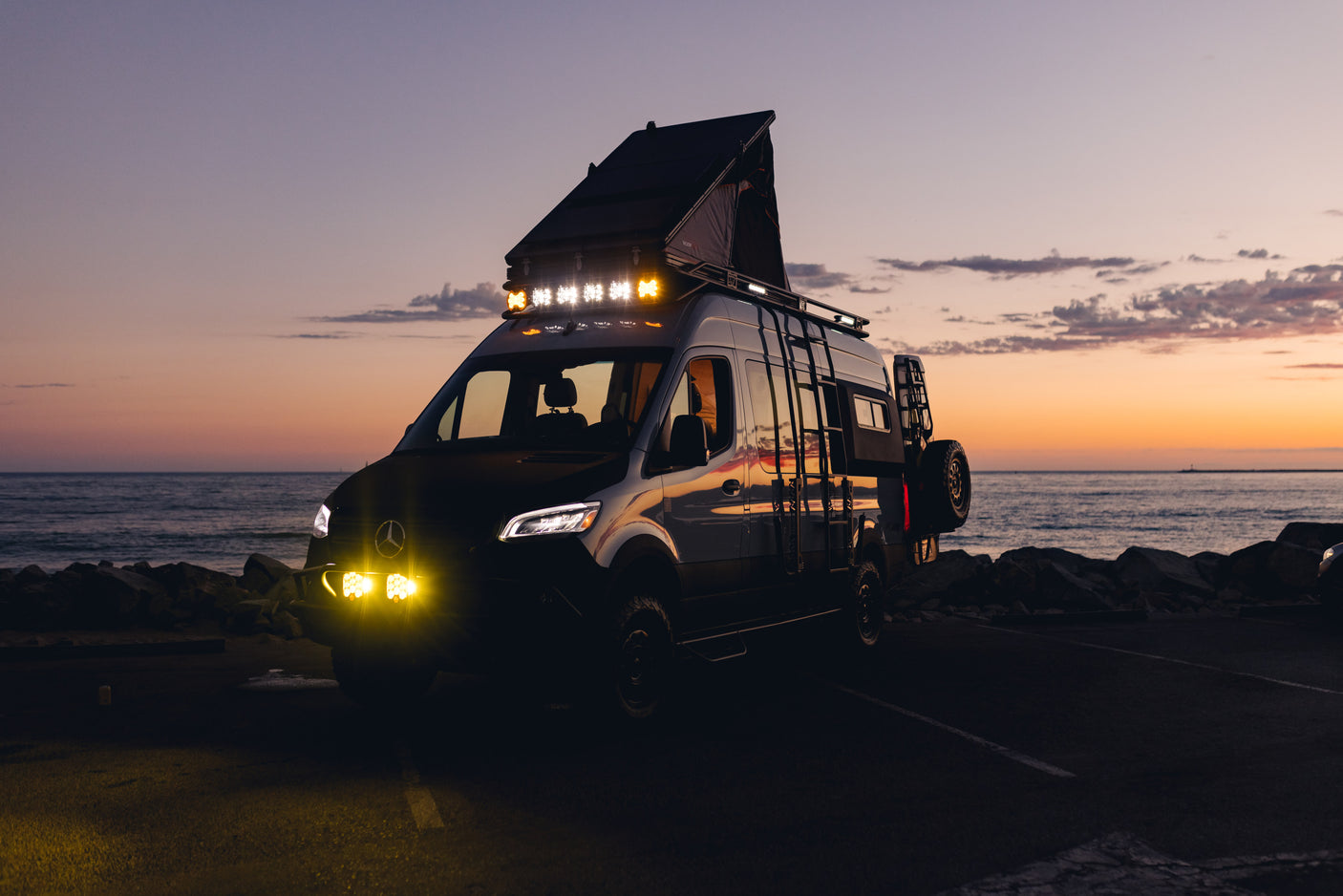 This adventure-ready Mercedes-Benz Sprinter RV rental, with a pop-top tent & bright lights, shines at sunset by the ocean.