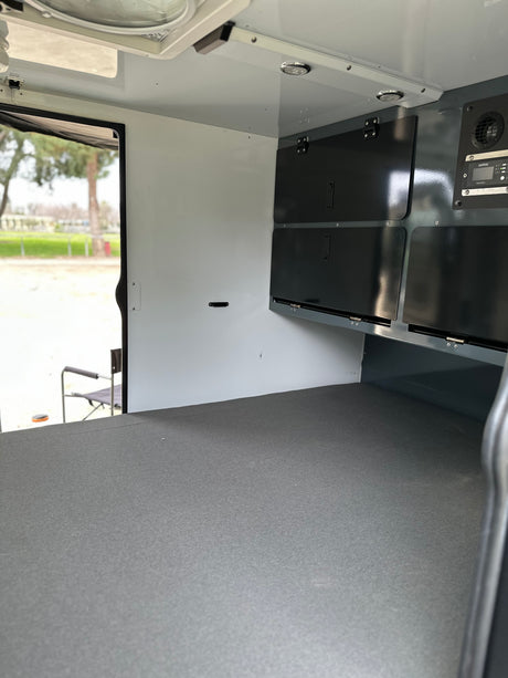 Inside an RV rental, featuring a grey sleeping platform, dark overhead storage, and a Xantrex electrical panel. Outdoor view.