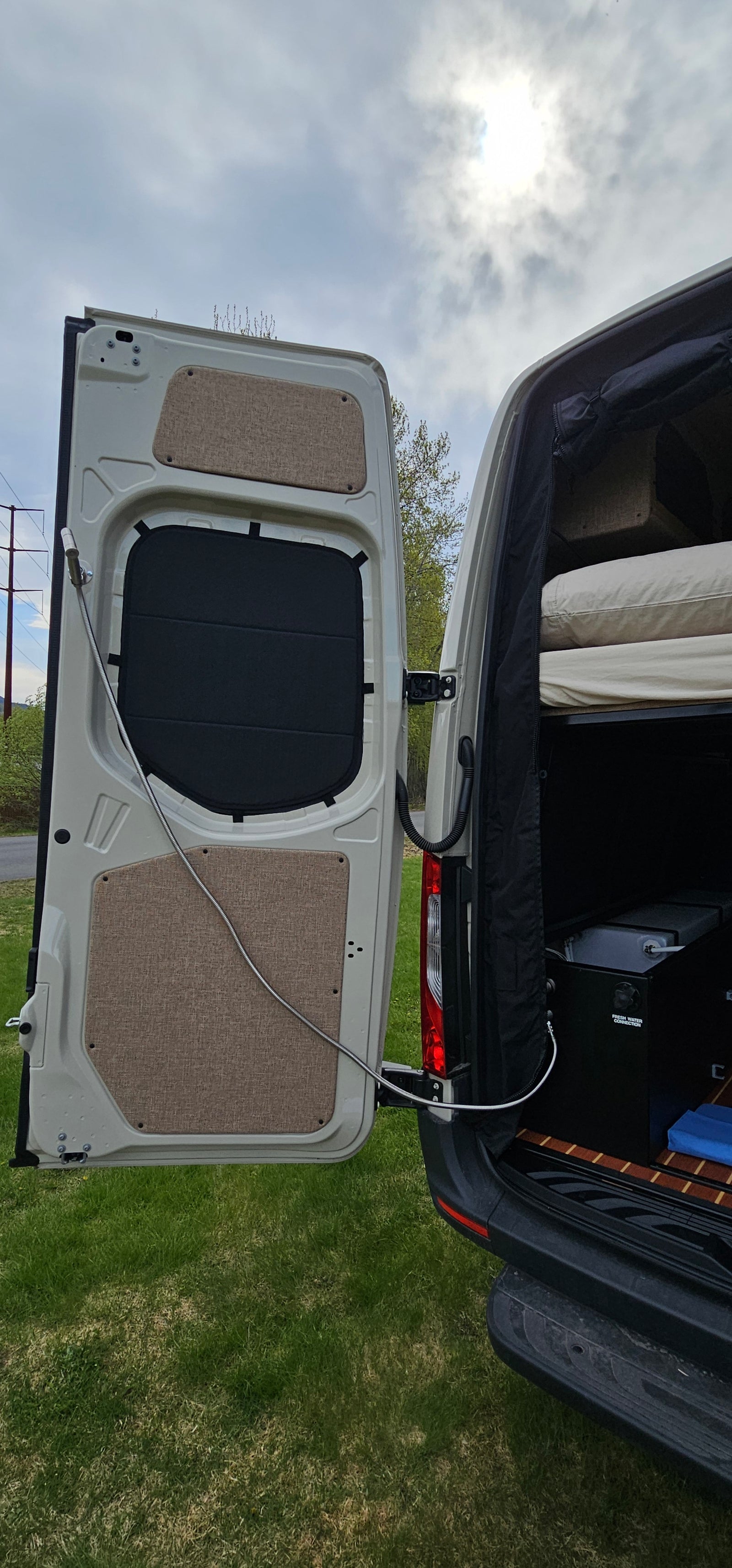 Open rear door of an RV rental showing bed, outdoor shower, and fresh water connection for mobile living.