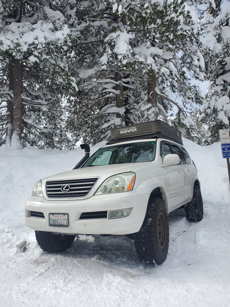 A white Lexus GX 470 with a rooftop tent is parked in deep snow among pine trees, ready for an rv rental outdoor adventure.