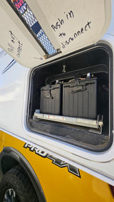 RV rental's battery compartment open, showing two batteries on a pull-out tray. Lid has handwritten instructions for connecting/disconnecting power.