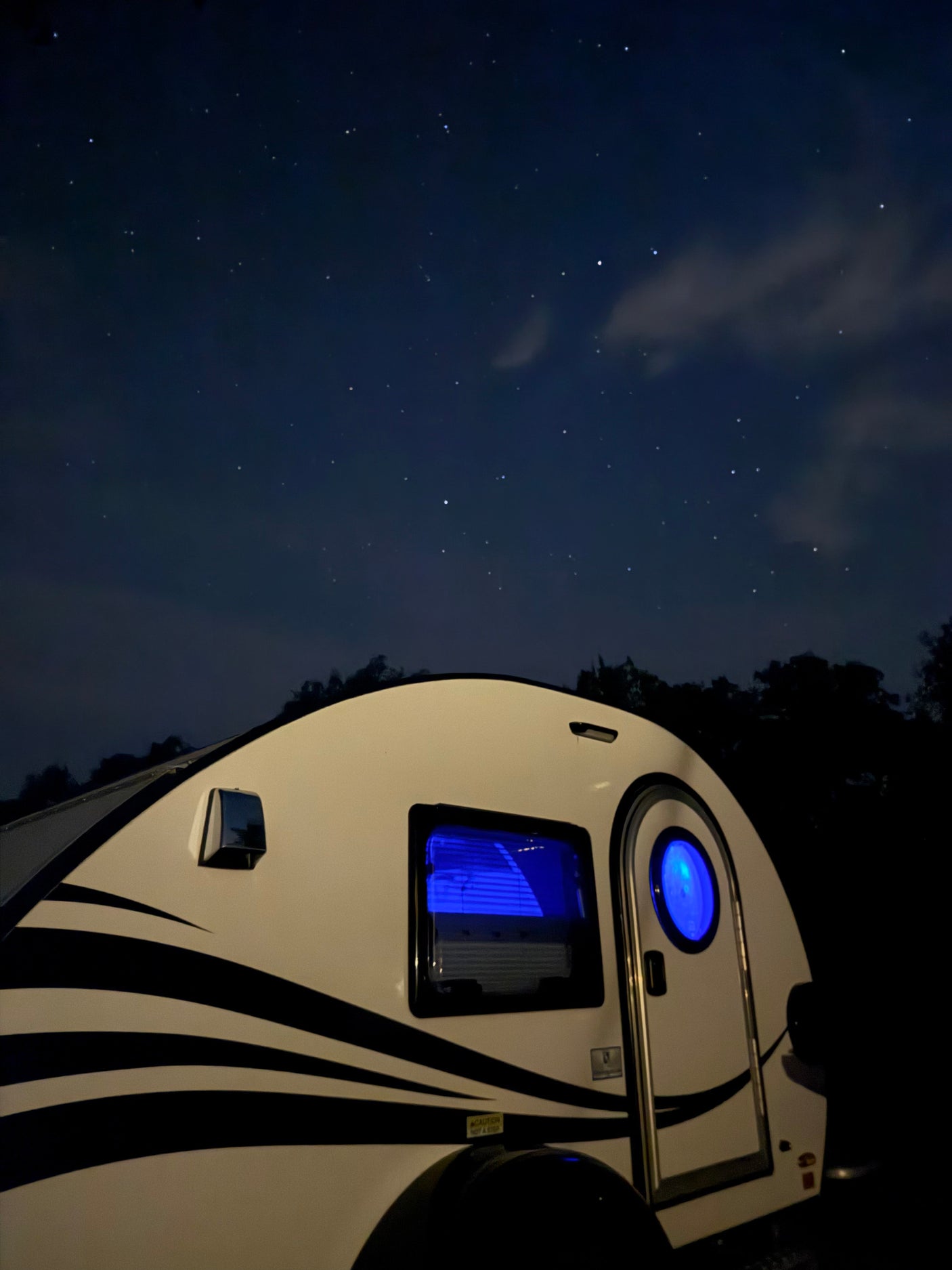 A cozy white teardrop **RV rental** glows blue under a stunning starry night sky, ideal for camping adventures.