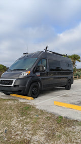 Dark gray Ram ProMaster camper van, likely an RV rental, parked under a cloudy sky with a bird perched on its roof.
